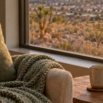 Sunlit living room chair with a knitted blanket, a mug of tea and desert wildflowers beside a window overlooking the Las Vegas valley at golden hour.